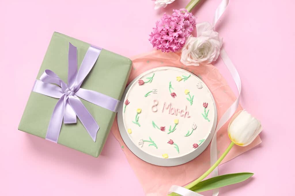Floral-themed bento cake with "8 March" written on top, beside a green gift box and spring flowers