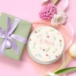 Floral-themed bento cake with "8 March" written on top, beside a green gift box and spring flowers