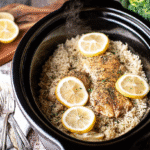 Slow cooker lemon pepper chicken with rice topped with lemon slices, served in a black crockpot with steam rising, surrounded by fresh lemons and broccoli.
