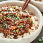 Slow cooker teriyaki chicken served over white rice, topped with sesame seeds and chopped green onions in a ceramic bowl.